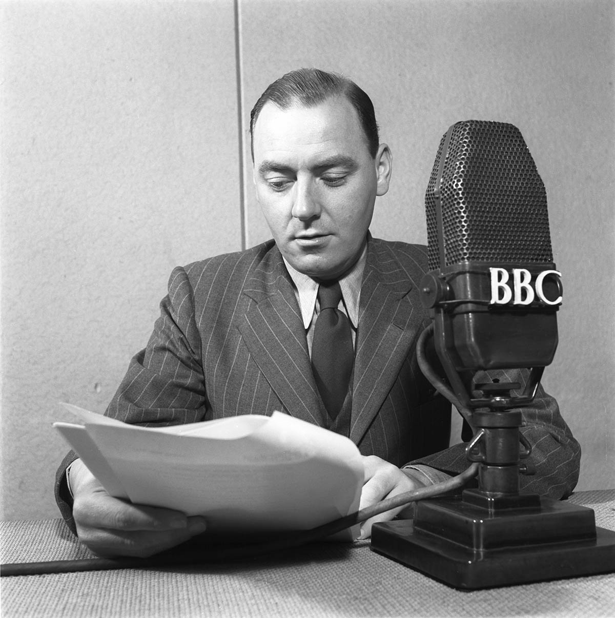 Frank Gillard, BBC war correspondent is seated in front of a large BBC microphone, holding some papers.