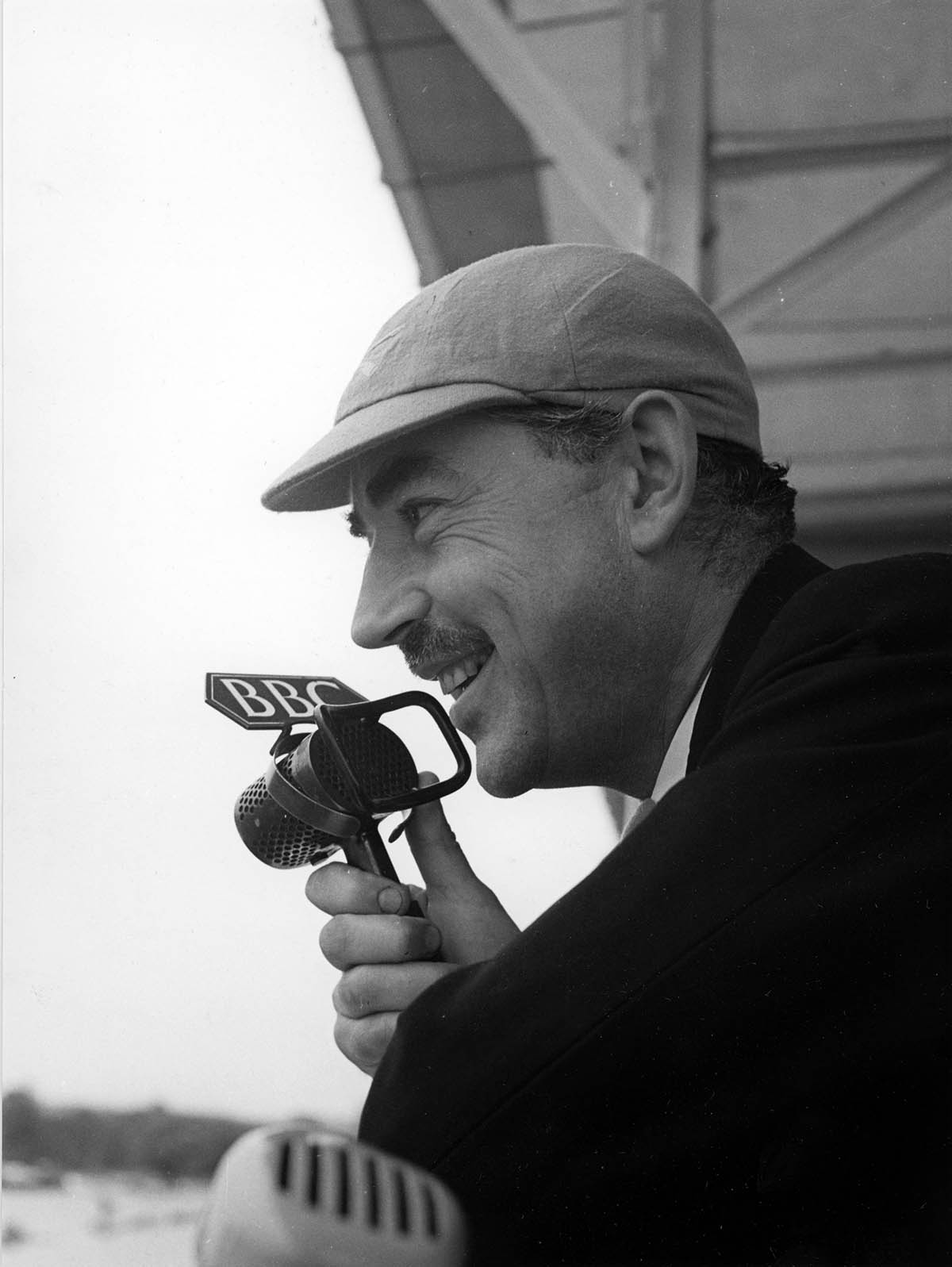 John Snagg, announcer, is pictured at a sporting event, holding a BBC microphone. This is a profile image, and he is wearing a peaked cap.